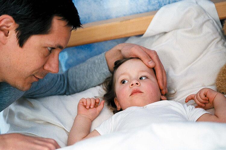 Kleines Mädchen liegt krank im Bett, der Vater legt ihr liebvevoll die Hand auf den Kopf.