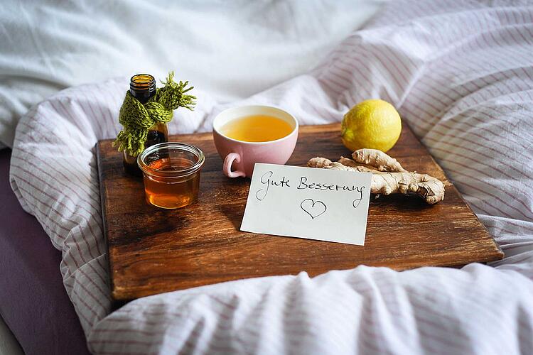 Hausmittel gegen Erkältung: Honig, Ingwer, Zitrone und Tee auf einem Holztablett auf einer Bettdecke