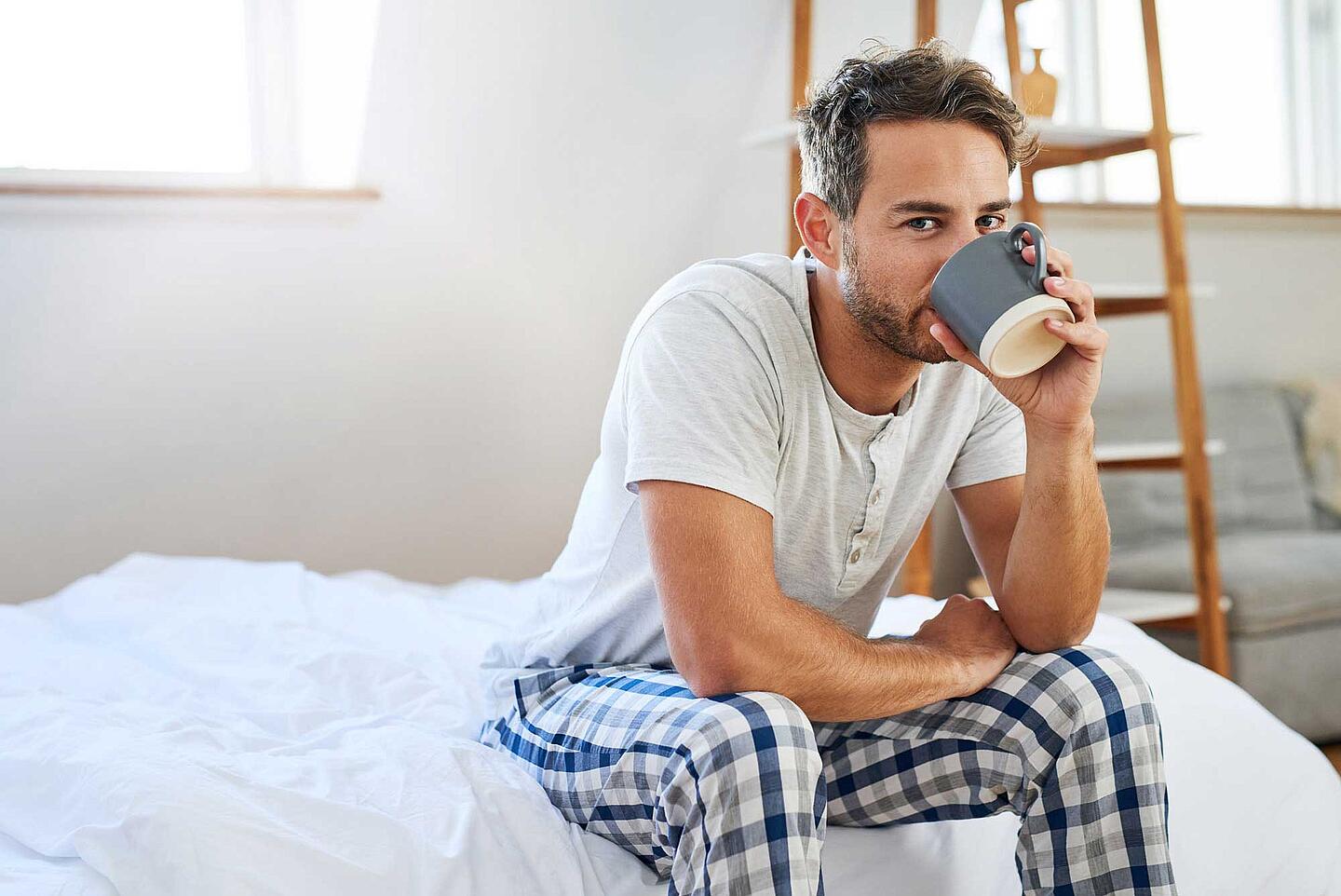Mann sitzt müde auf der Bettkante und trinkt Kaffee.