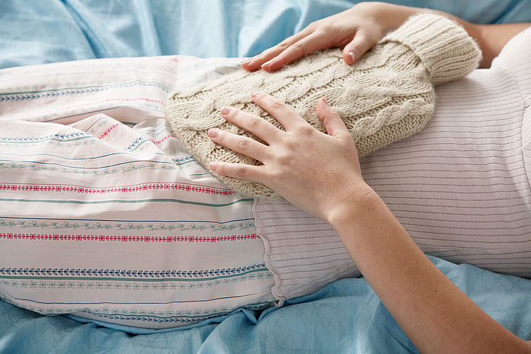 Bildnachweis: © wdv / Oana Szekely Frau mit Wärmflasche auf dem Bauch liegt auf dem Bett.