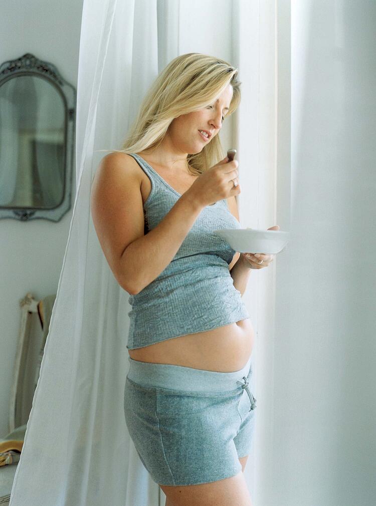 Schwangere Frau steht am Fenster und isst.