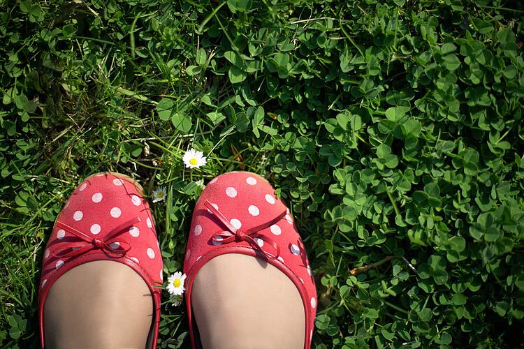 Füße in roten Ballerinas mit weißen Punkten auf einer Wiese mit Gänseblümchen.