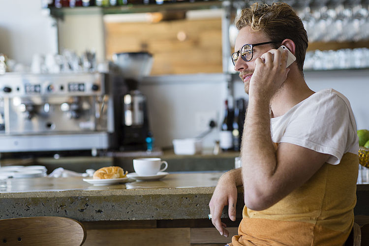 Ein junger Mann frühstückt in einem Cafe und telefoniert mit seinem Handy.