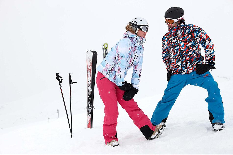 Zwei Alpin-Skifahrer machen Dehnungsübungen im Schnee.