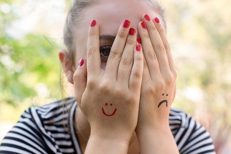 Eine junge Frau spickt durch die mit lachendem und traurigem Smiley bemalten Hände vor ihrem Gesicht.