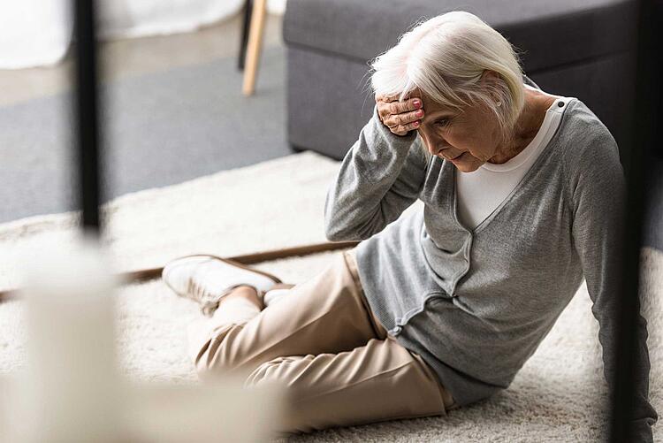 Seniorin sitzt am Boden und hält sich die Hand an die Stirn