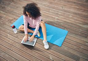 Frau sitzt auf einer Yogamatte und tippt auf einem Laptop
