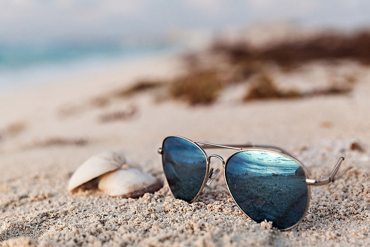 Bildnachweis: © istockphoto.com / Evgeniyaphotography Eine Sonnenbrille liegt neben zwei Muscheln im Sand.