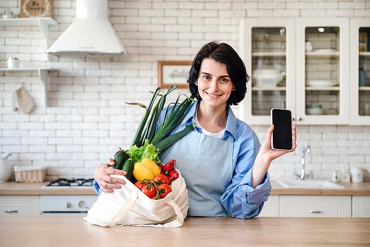 Fasten in der Fastenzeit: Frau mit Einkaufstasche voll mit frischem Gemüse hält ein Smartphone in der Hand