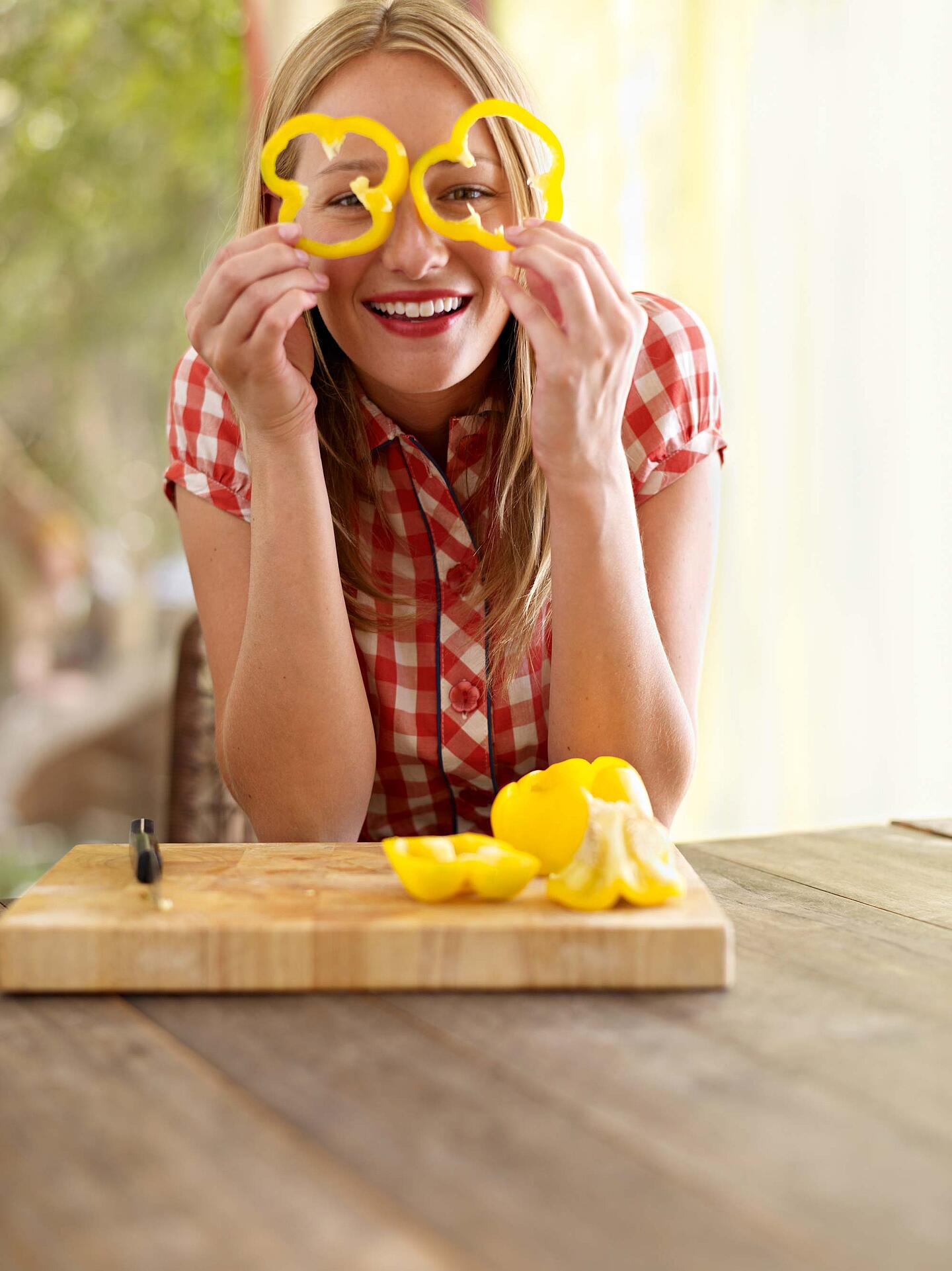 Eine junge, lachende Frau hält sich Paprikaringe wie eine Brille vor die Augen.