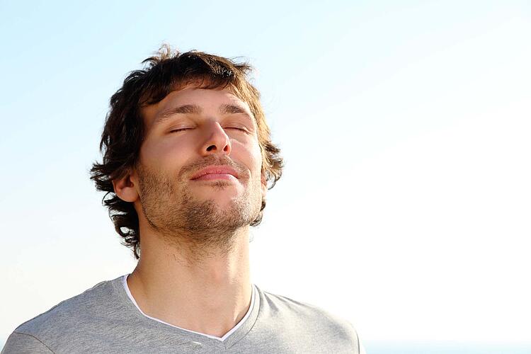 Junger Mann atmet mit geschlossenen Augen, im Hintergrund blauer Himmel.