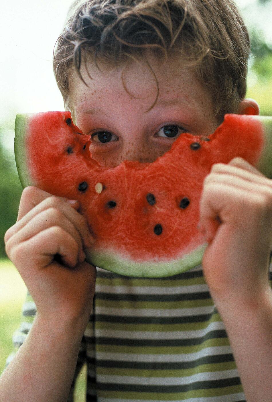 Ein Junge hält ein großes Stück Wassermelone vor sein Gesicht.