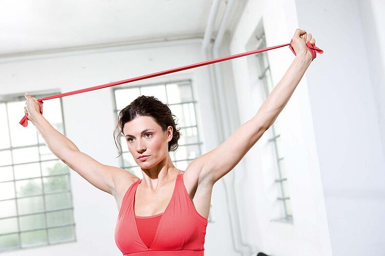Junge Frau in rotem Sporttop dehnt ein Theraband über dem Kopf.