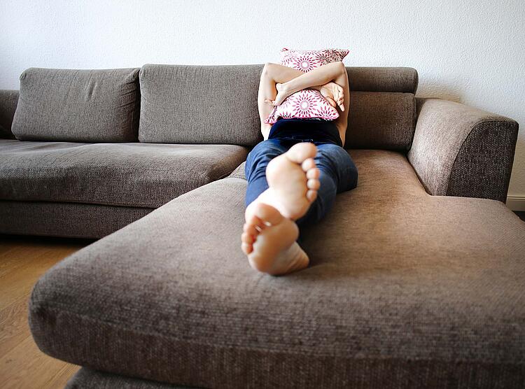 Junge Frau liegt mit nackten Füßen auf einem Sofa und hält ein Kissen vor das Gesicht.
