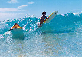 Zwei junge Frauen beim Wellenreiten auf Surfbrettern.