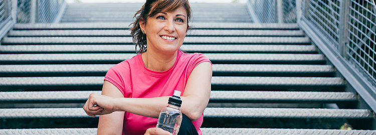 Eine Frau sitzt mit einer Trinkflasche in der Hand auf einer Treppe. Eine Frau sitzt mit einer Trinkflasche in der Hand auf einer Treppe.