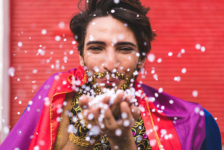 Mann in einem Karnevalskostüm mit goldenem Glitzer im Bart pustet in seine Hände voll mit Konfetti.