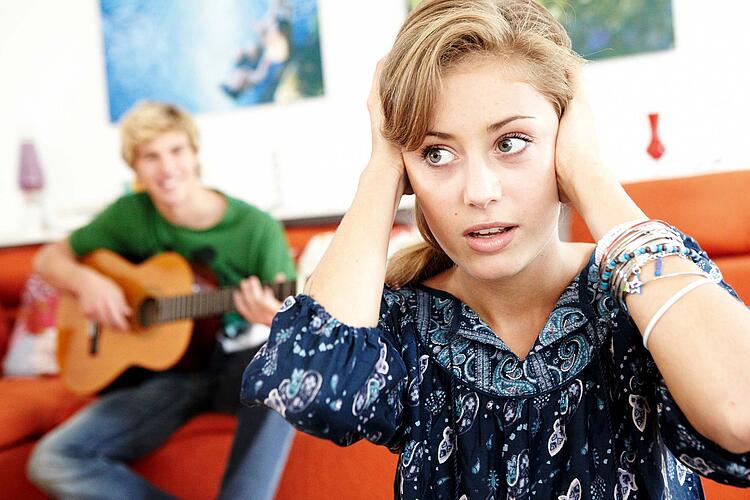 Junge Frau hält sich gestresst die Ohren zu während ein junger Mann im Hintergrund Gitarre spielt.