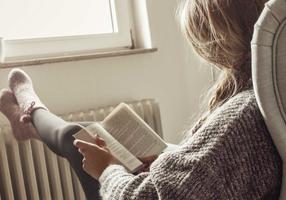 Frau sitzt im Sessel mit den Füßen auf der Heizung und liest
