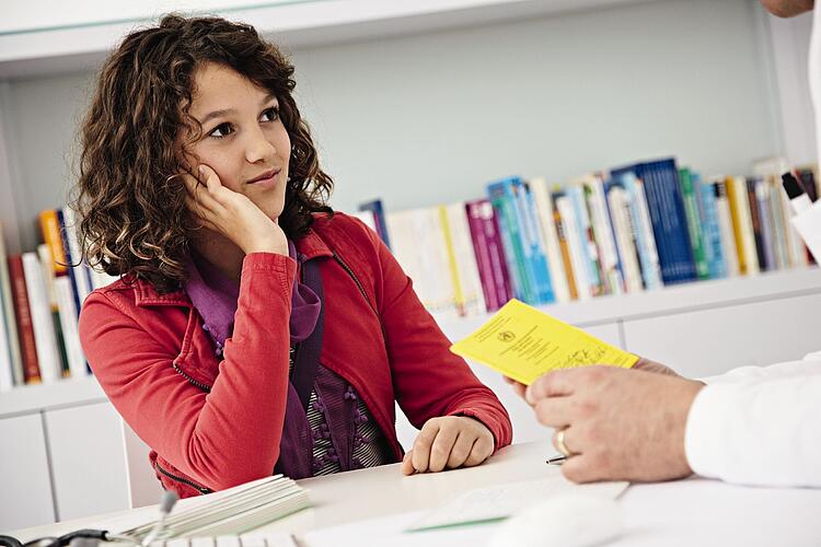 Junge Frau beim Arzt, der Arzt hält einen Impfpass in der Hand.