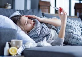 Frau liegt krank auf dem Sofa und misst Fieber