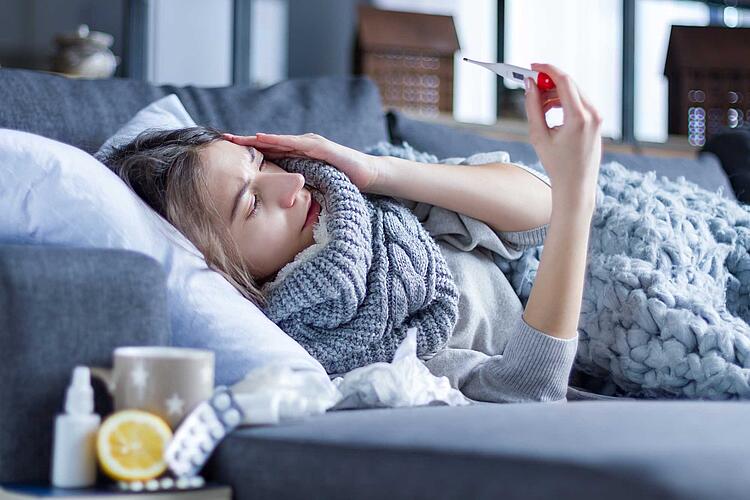 Frau liegt krank auf dem Sofa und misst Fieber