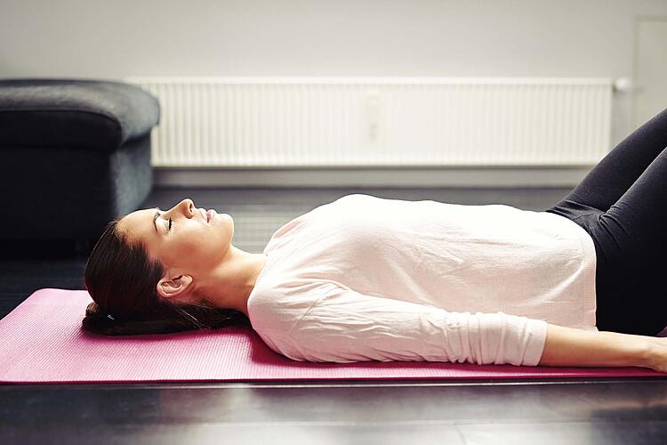 Junge Frau liegt mit geschlossenen Augen und angezogenen Beinen auf einer Yogamatte.