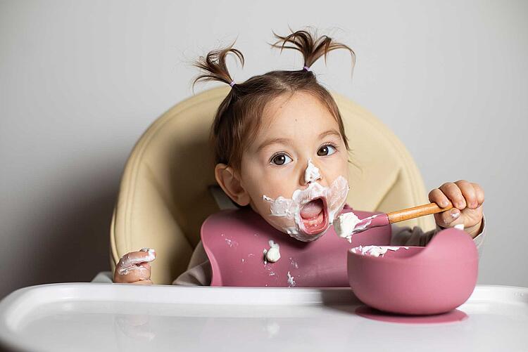 Kleines Mädchen im Hochstuhl ist Joghurt mit einem Löffel, das ganze Gesicht ist mit Joghurt verschmiert.