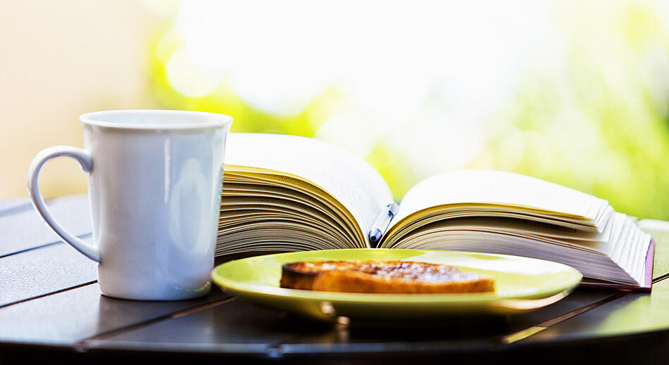 Eine Scheibe Toast auf einem Teller und eine Tasse stehen vor einem aufgeschlagenen Buch.