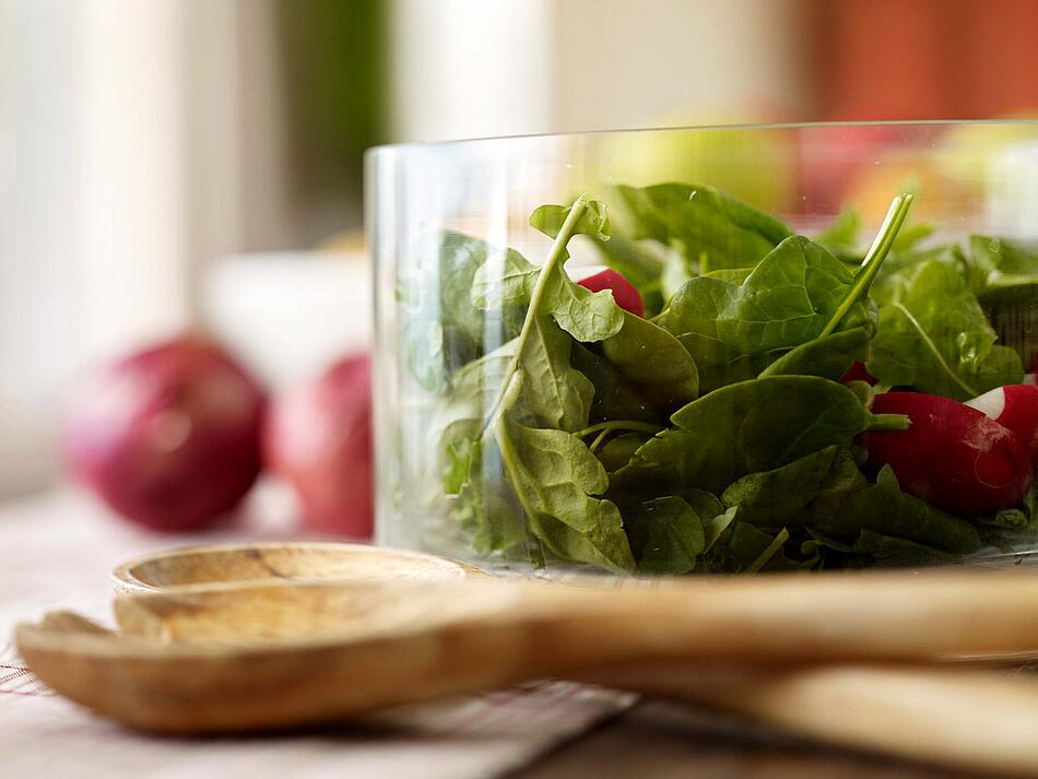 Grüner Salat und Radieschen in einer Glasschüssel mit einem Salatbesteck aus Holz im Vordergrund.