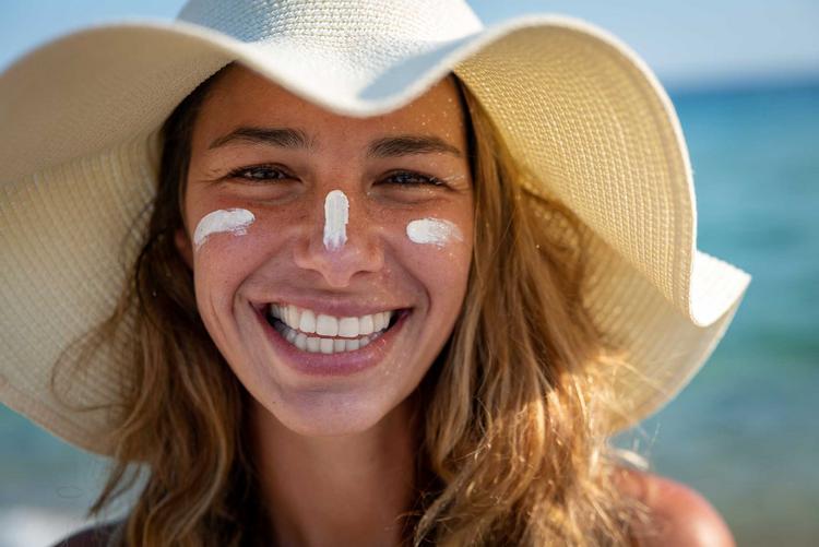 Junge Frau mit Sonnenhut und Sonnencreme im Gesicht strahlt in die Kamera