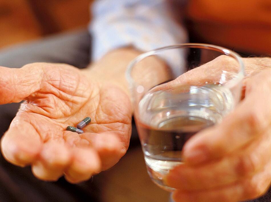 Männerhände, in der einen Hand liegen Tabletten, die andere Hand hält ein Glas Wasser.