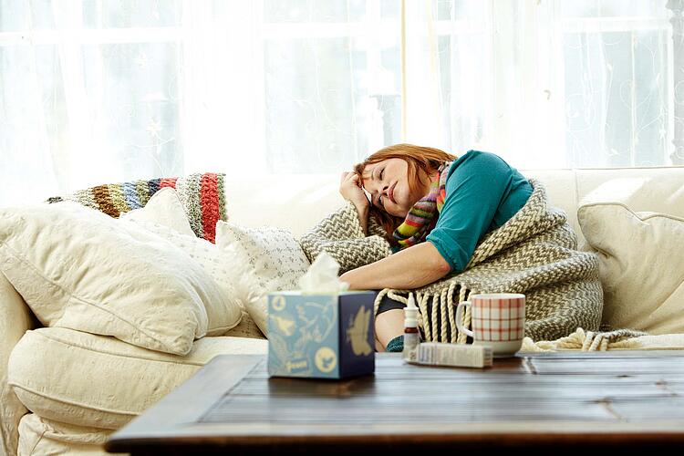 Eine erkältete Frau sitzt in eine Wolldecke gewickelt auf dem Sofa.
