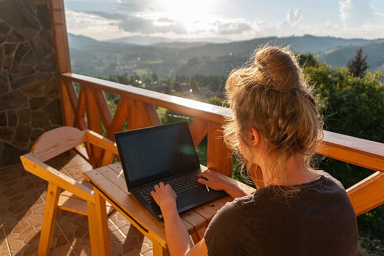 Workation – Frau arbeitet am Laptop auf einem Balkon mit Blick über ein Tal