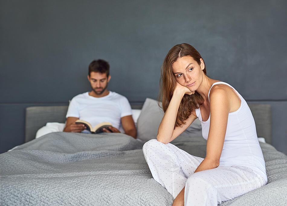 Paar im Schlafzimmer, Mann liest, Frau sieht genervt aus
