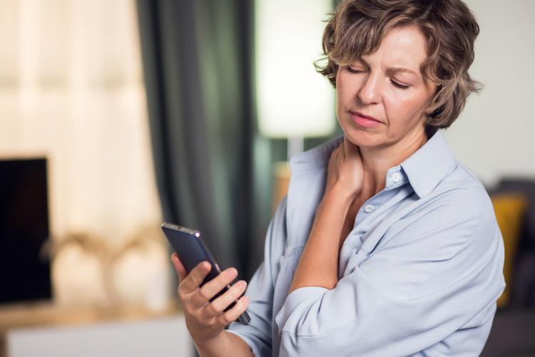 Frau mit Smartsphone in der Hand greift sich an den schmerzenden Nacken