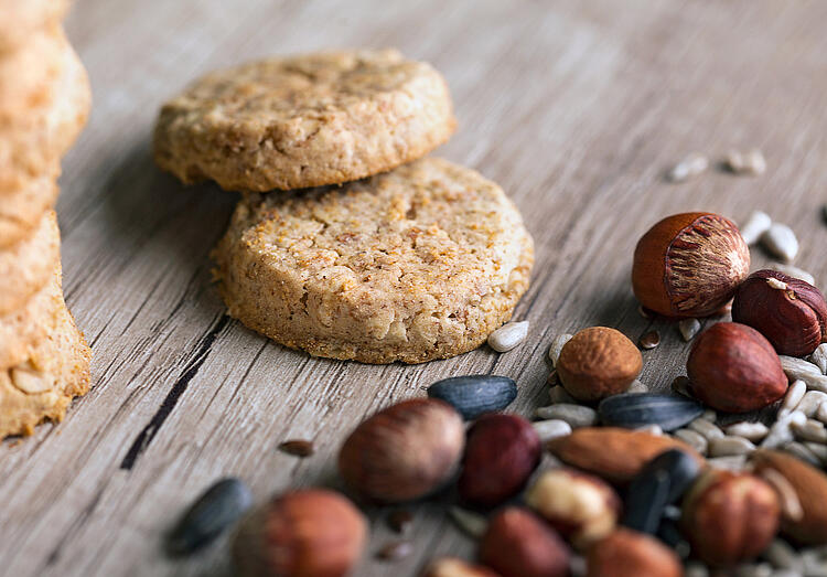 Zwei Nusskekse liegen auf einem Holztisch, daneben Haselnüsse, Mandeln, Kürbiskerne und Sonnenblumenkerne. 