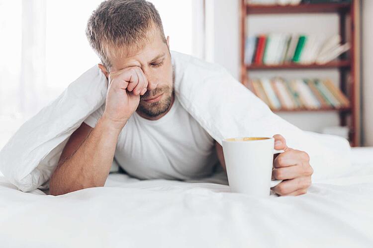 Verschlafen wirkender Mann trinkt im Bett eine Tasse Kaffee
