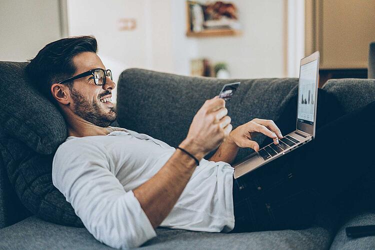 Mann liegt bequem auf der Couch und kauft am Laptop online ein.