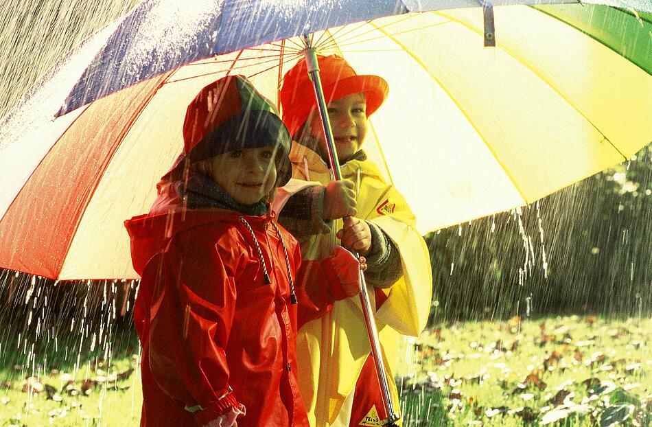 Zwei Kinder in Regenkleidung unter einem Schirm.