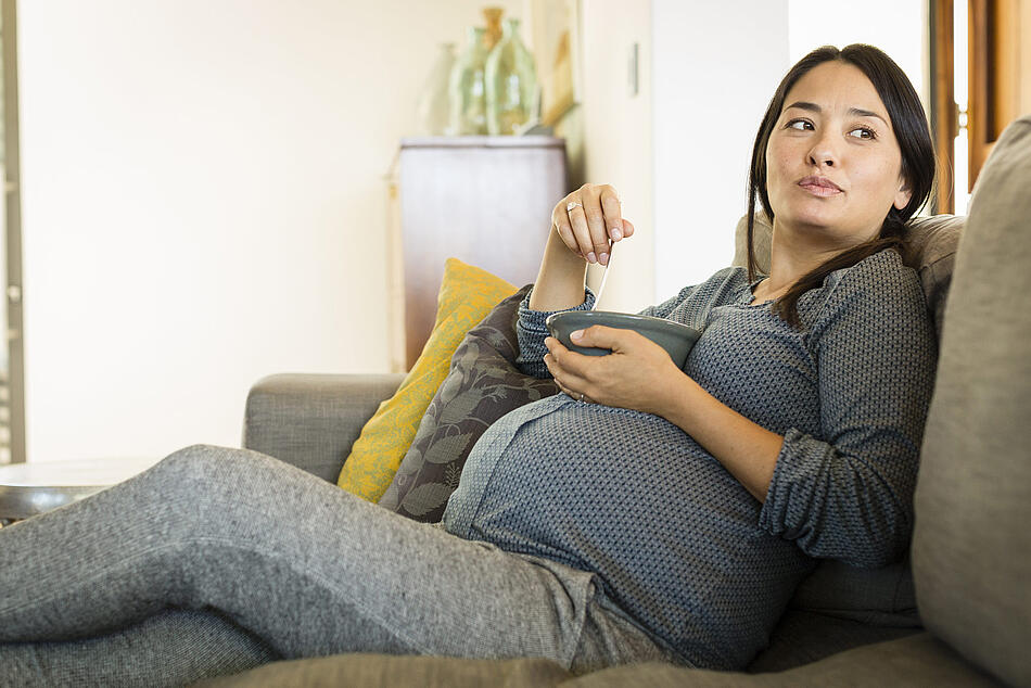 Schwangere sitzt auf dem Sofa und isst aus einer Schüssel in ihrer Hand.