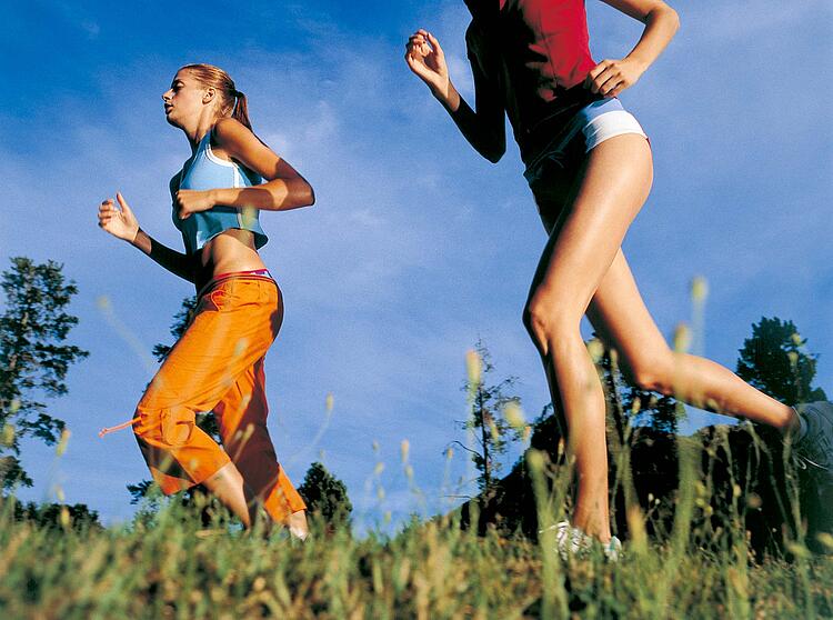 Zwei Frauen joggen in sommerlicher Sportkleidung über eine Wiese, blauer Himmel im Hintergrund.