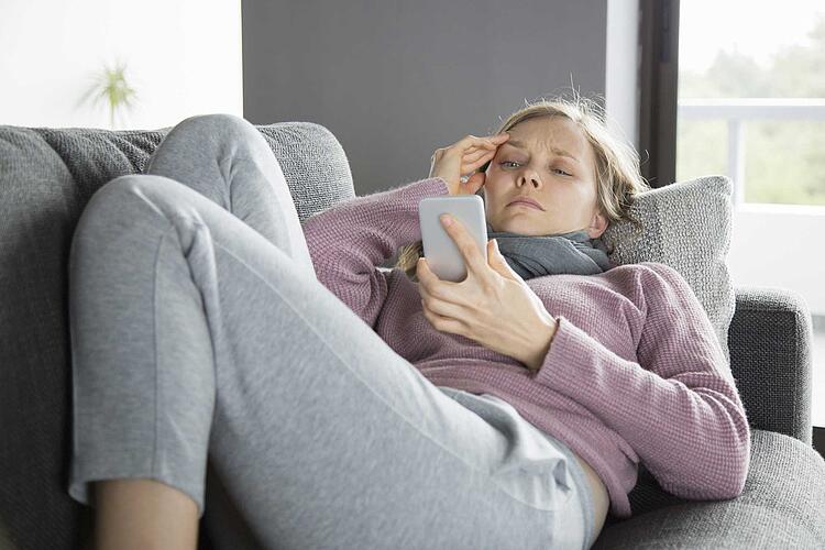 Frau liegt krank auf dem Sofa und hat ein Smartphone in der Hand.