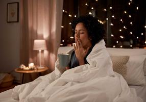 Frau sitzt im Bett, trinkt eine Tasse Tee und gähnt