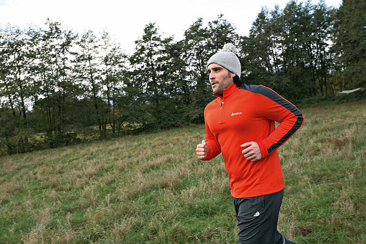 Junger Mann in Sportkleidung mit Strickmütze joggt über eine Wiese mit Bäumen im Hintergrund.