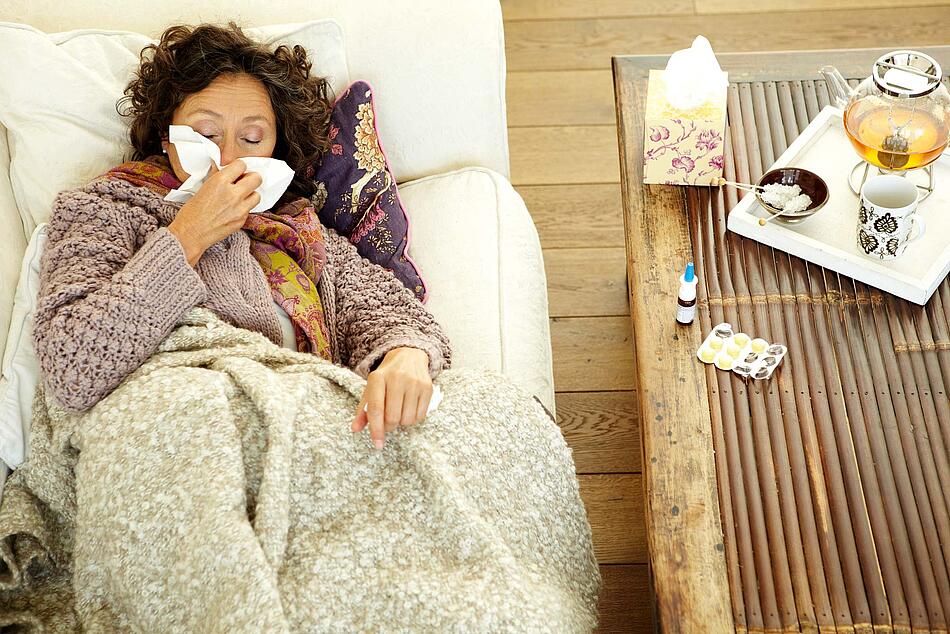 Erkältete Frau liegt zugedeckt auf dem Sofa und putzt sich die Nase.