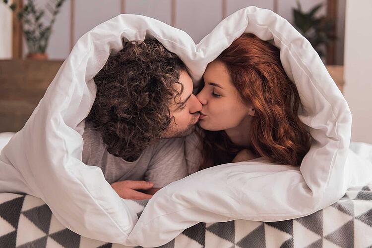 Junges Paar küsst sich unter der Bettdecke, die in Herzform um das Paar geschlungen ist.