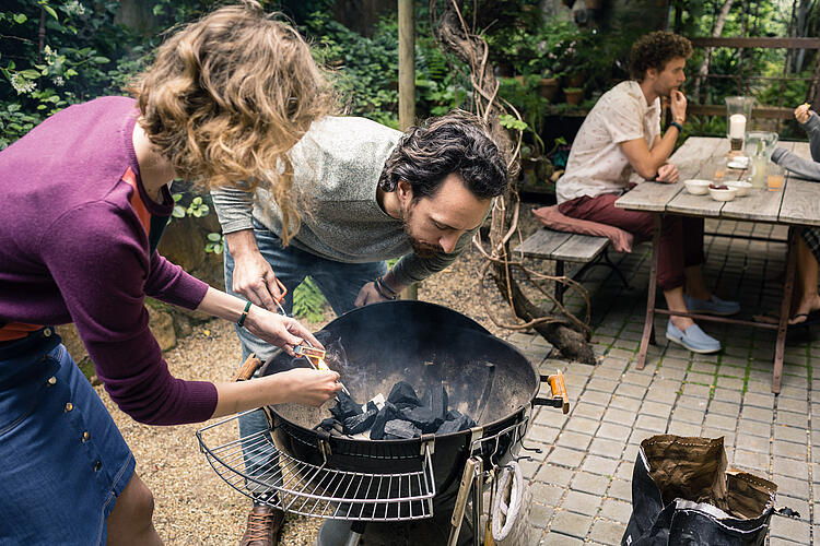 Ein junges Paar zündet gemeinsam einen Holzkohlegrill an, im Hintergrund sitzt ein Mann an einer Bierzeltgarnitur.