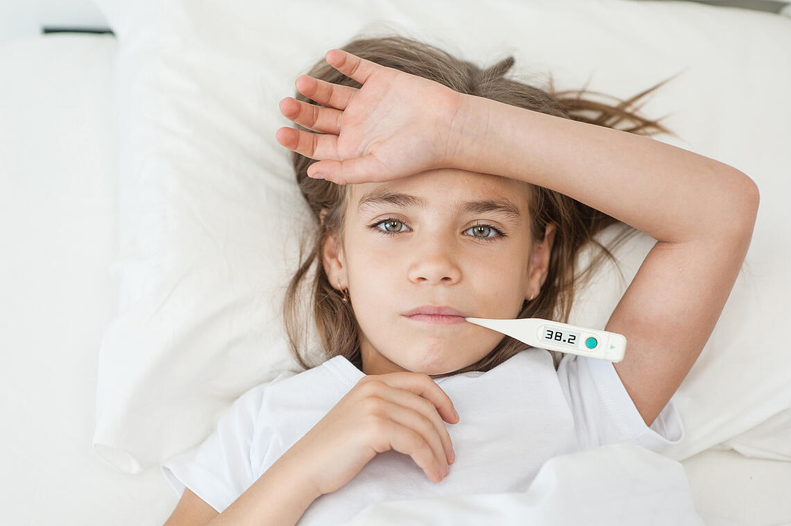 Mädchen liegt mit einem Fieberthermometer im Mund im Bett.