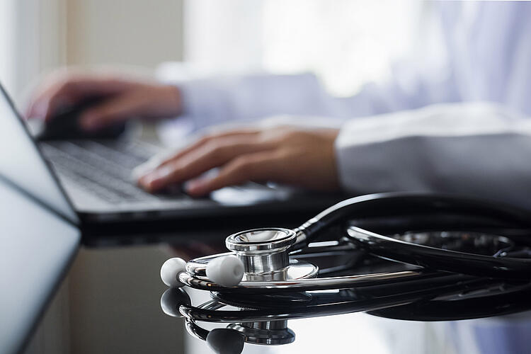 Arzt tippt auf einem Laptop, im Vordergrund ein Stethoskop
